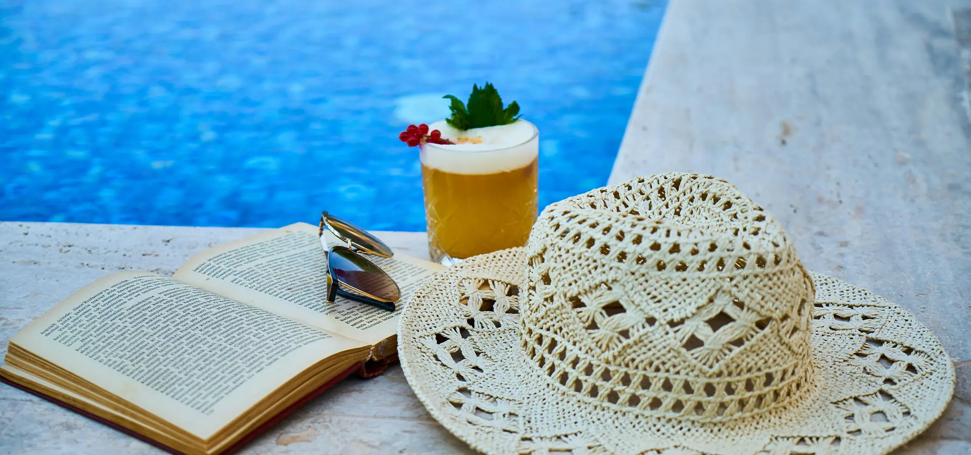 Strohhut, Buch, Sonnenbrille und Getränk am Beckenrand