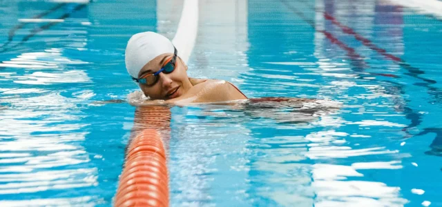 Schwimmerin mit Taucherbrille und Bademütze lehtn sich auf ein Bahnenseil im Wasser