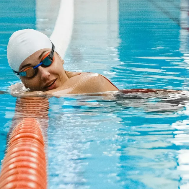 Schwimmerin mit Taucherbrille und Bademütze lehtn sich auf ein Bahnenseil im Wasser